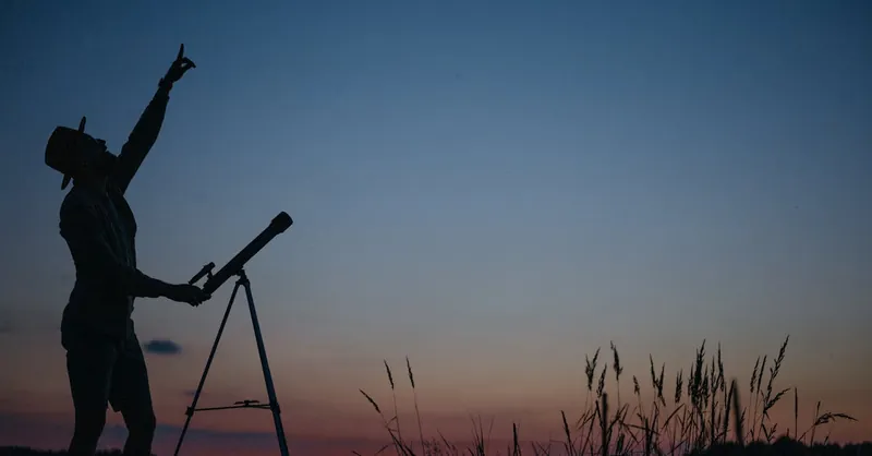Silhouette of a person pointing at the sky with a telescope during twilight.