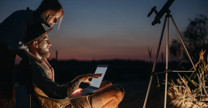 A couple using a laptop and telescope under the night sky, enjoying stargazing outdoors.