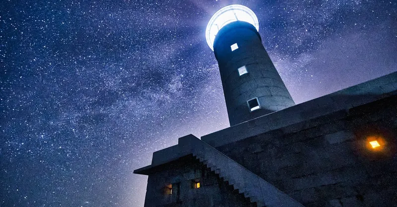 Low-angle view of a lighthouse illuminated against a starry night sky, creating a dramatic celestial scene.