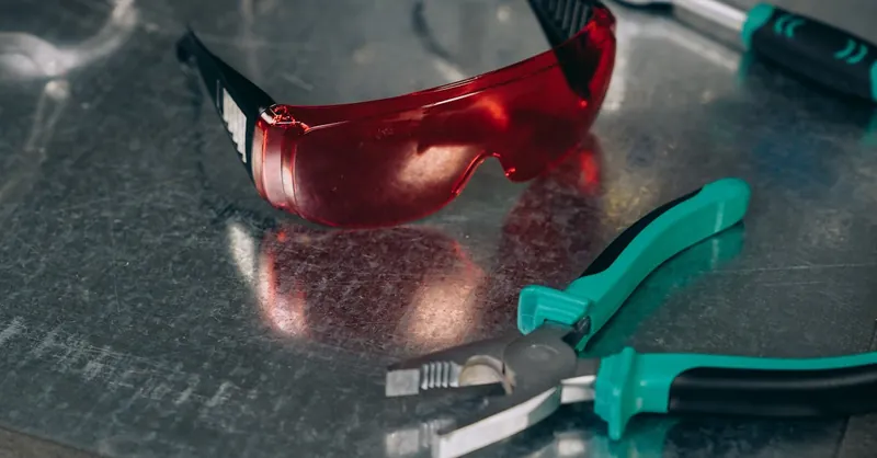 Red safety goggles and green-handled pliers on a reflective metal surface, emphasizing protective tools.