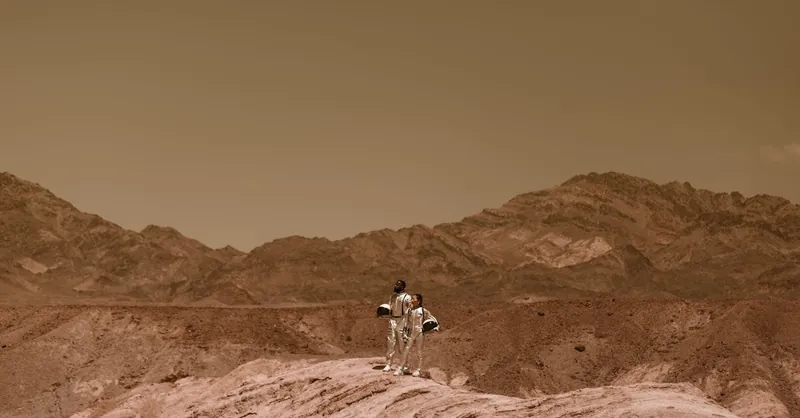 Two astronauts in space suits explore a Mars-like rocky desert landscape under a warm, sepia sky.