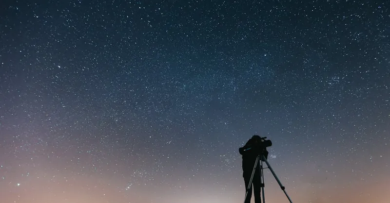 A silhouetted person using a telescope to observe the night sky filled with stars.