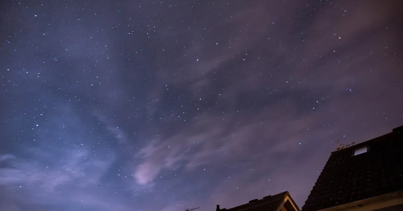 Long-exposure capture of a star-filled night sky with clouds and rooftops in view.